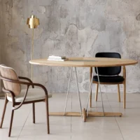 A modern dining area with a round wooden table, two different chairs (one wood and one black), a gold floor lamp, and stacked books, set against a textured concrete wall.
