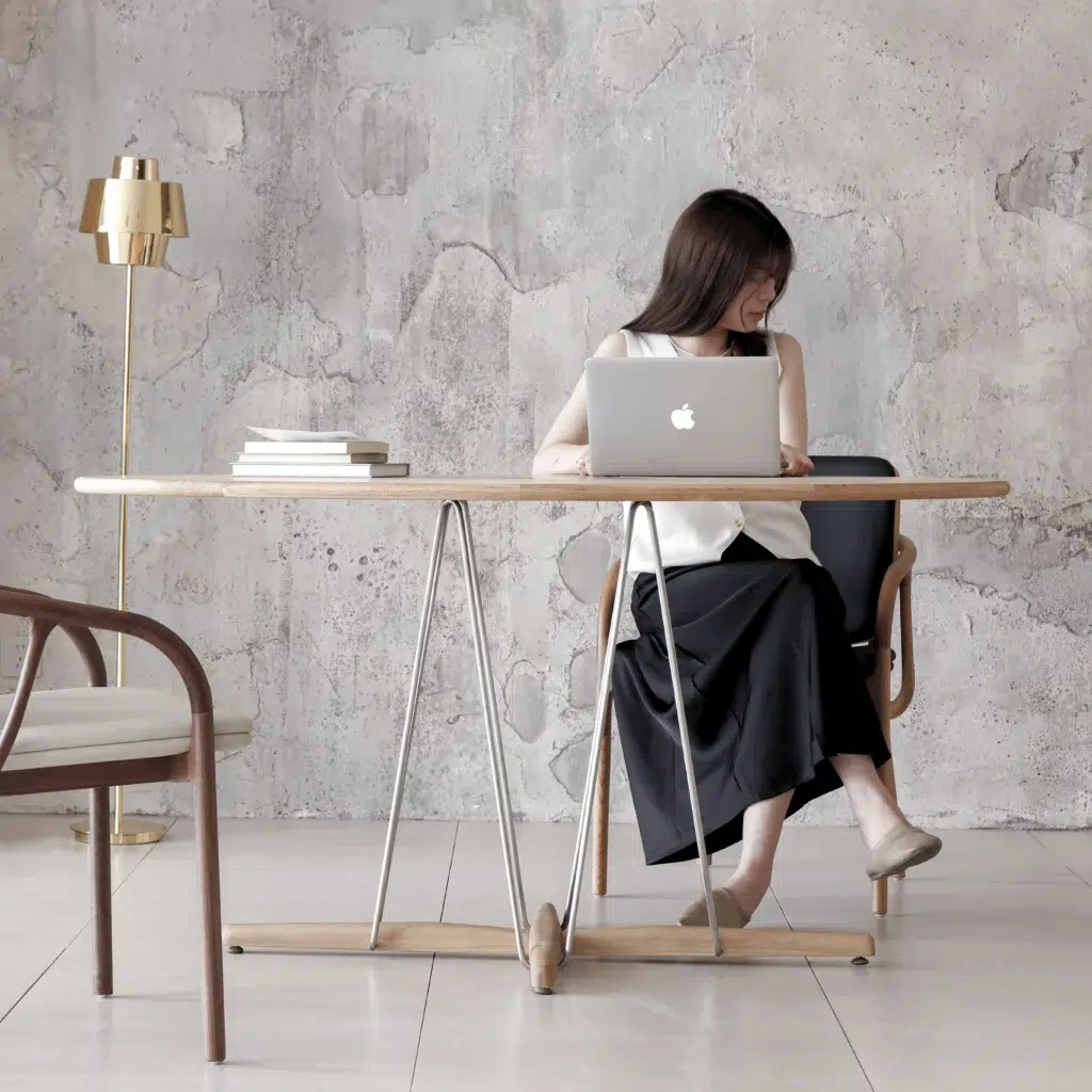 A woman sits at a wooden table with a laptop, looking to her side. The setting is minimalistic, with modern furniture, a stack of books, and a gold floor lamp against a textured, light grey wall.