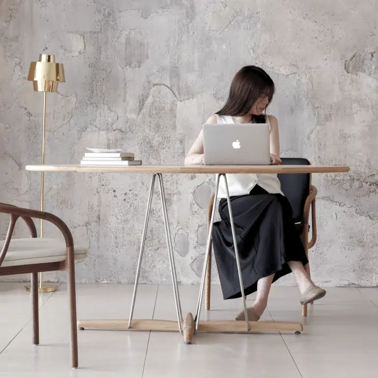 A woman sits at a wooden table with a laptop, looking to her side. The setting is minimalistic, with modern furniture, a stack of books, and a gold floor lamp against a textured, light grey wall.