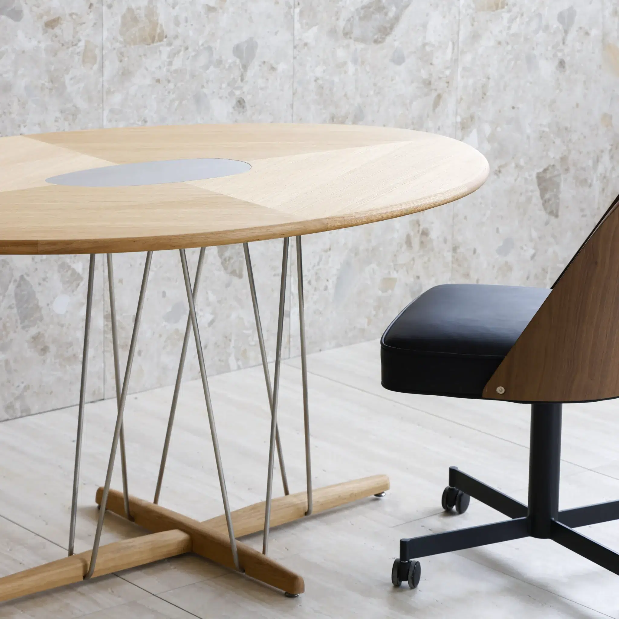 A modern wooden round table with thin metal legs stands next to a black and wood office chair with castors, set against a light stone wall and light-coloured floor.