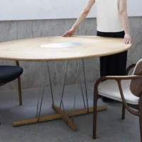A person stands behind a large, round wooden table with metal legs, touching its edge. Two modern chairs, one black and one white, are positioned on either side of the table in a minimalistic room.