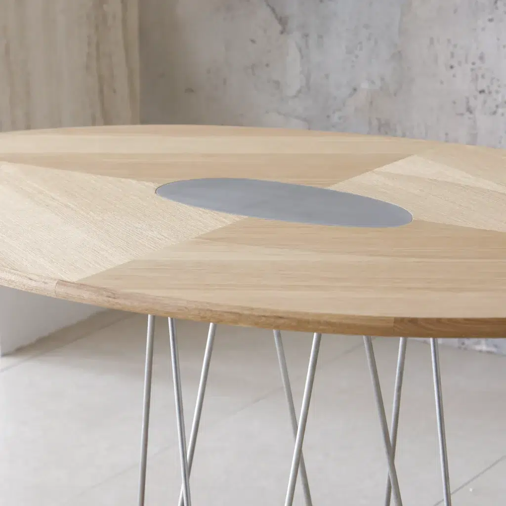 A modern wooden table with a geometric pattern and a grey oval centrepiece, supported by thin, angled metal legs, set against a textured light-coloured wall and floor.