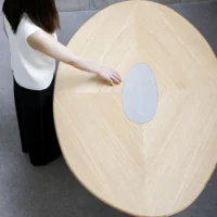 A person with long dark hair, wearing a white top and black trousers, stands beside a large oval wooden table with a light wood grain and an oval metallic centrepiece, touching the surface.