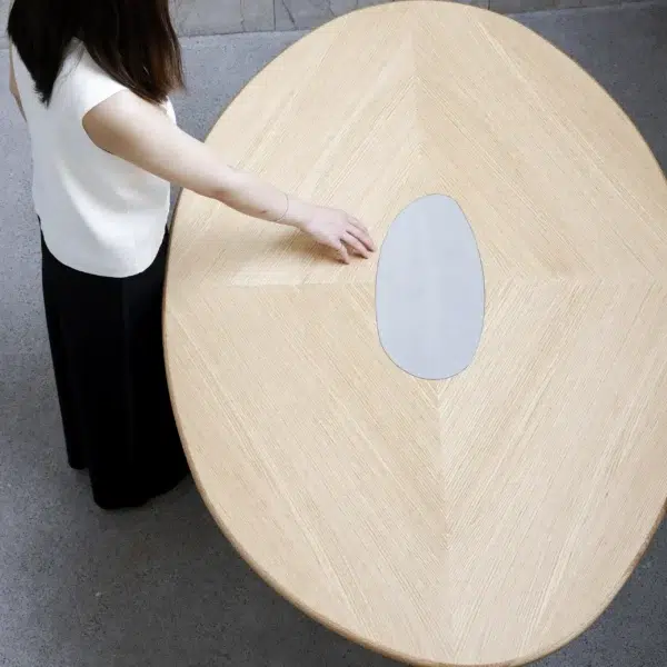 A person with long dark hair, wearing a white top and black trousers, stands beside a large oval wooden table with a light wood grain and an oval metallic centrepiece, touching the surface.