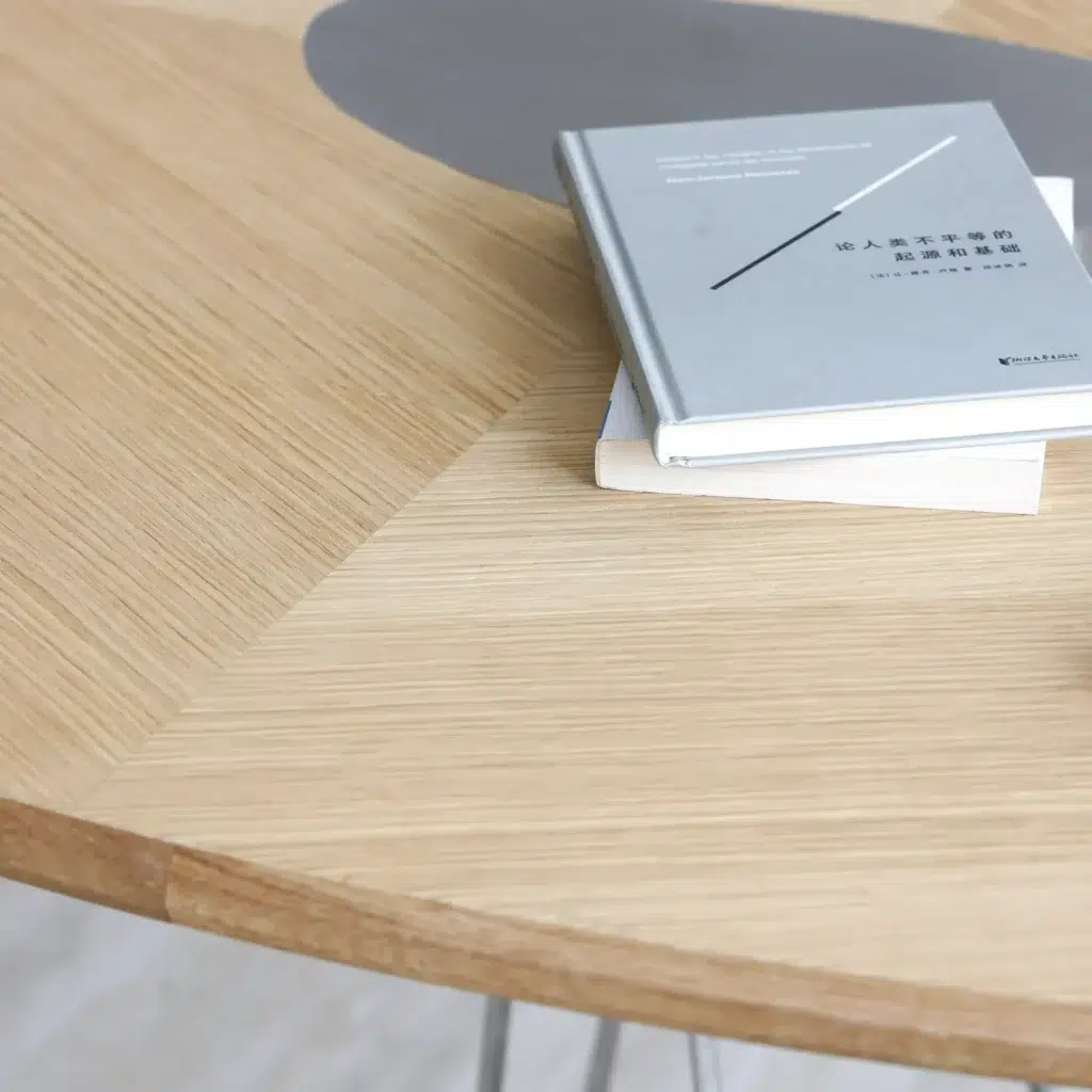 A stack of two closed books rests on a round, light wood table with a geometric pattern and a grey accent. The top book has a minimalist grey cover with Chinese text and a thin diagonal line design.