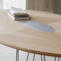 A light wooden table with a grey oval inset in the centre, holding a small stack of books and a notebook with handwritten notes, in a bright, modern room.