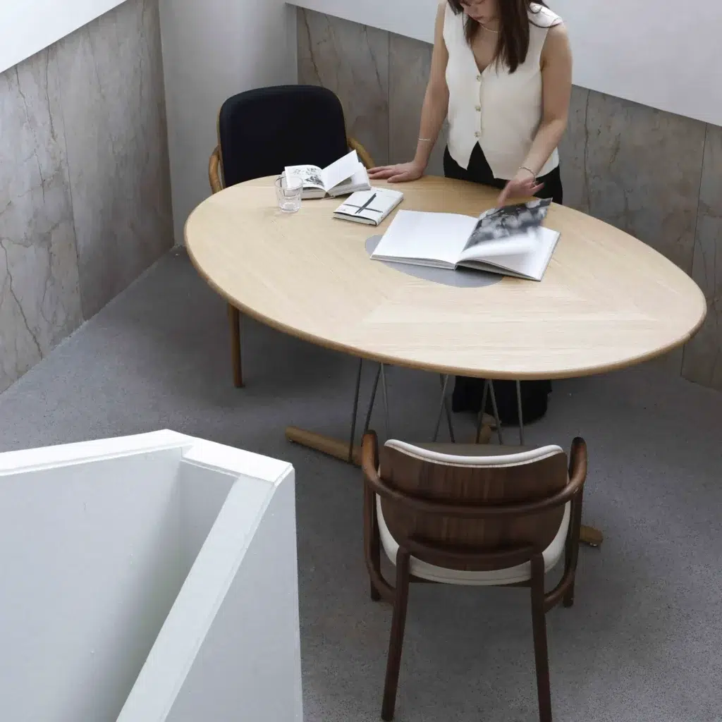 A woman stands at an oval wooden table, looking through open books. Two chairs are positioned at the table, one empty and one partially visible. The setting is minimalistic with light walls and carpeting.