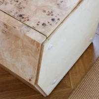 A close-up of a wooden box with a light wood grain and pattern, featuring rounded edges and white circular pegs on the side. The box sits on a wooden floor next to a woven beige rug.
