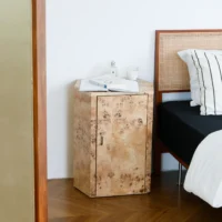 A light wood bedside table with a unique grain pattern stands beside a bed with a woven rattan headboard. The bedside table has an open book, a candle, and a small bottle on top. The bed has black and white striped bedlinen.