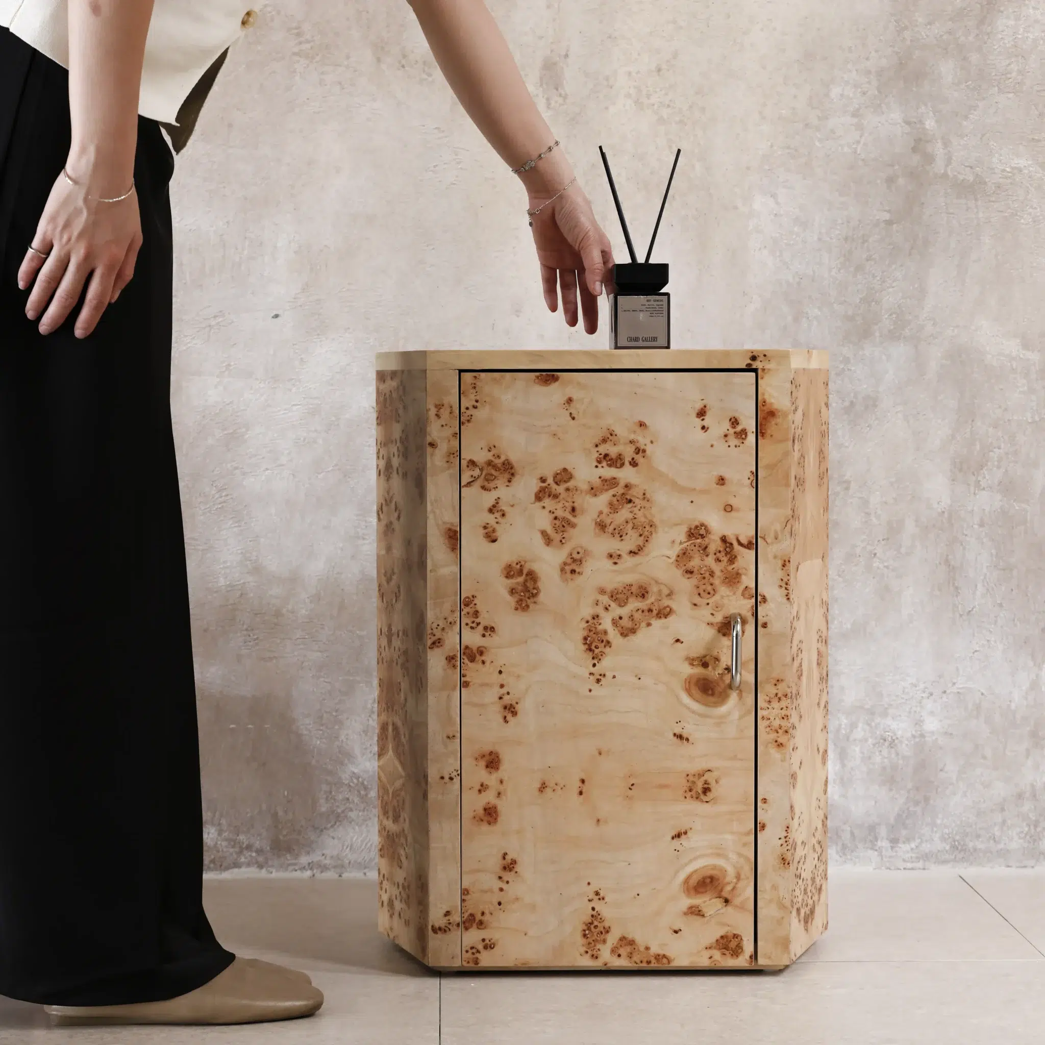 A person reaches to place a fragrance diffuser on a small, hexagonal wooden cabinet with a natural, burr wood pattern, set against a textured beige wall.