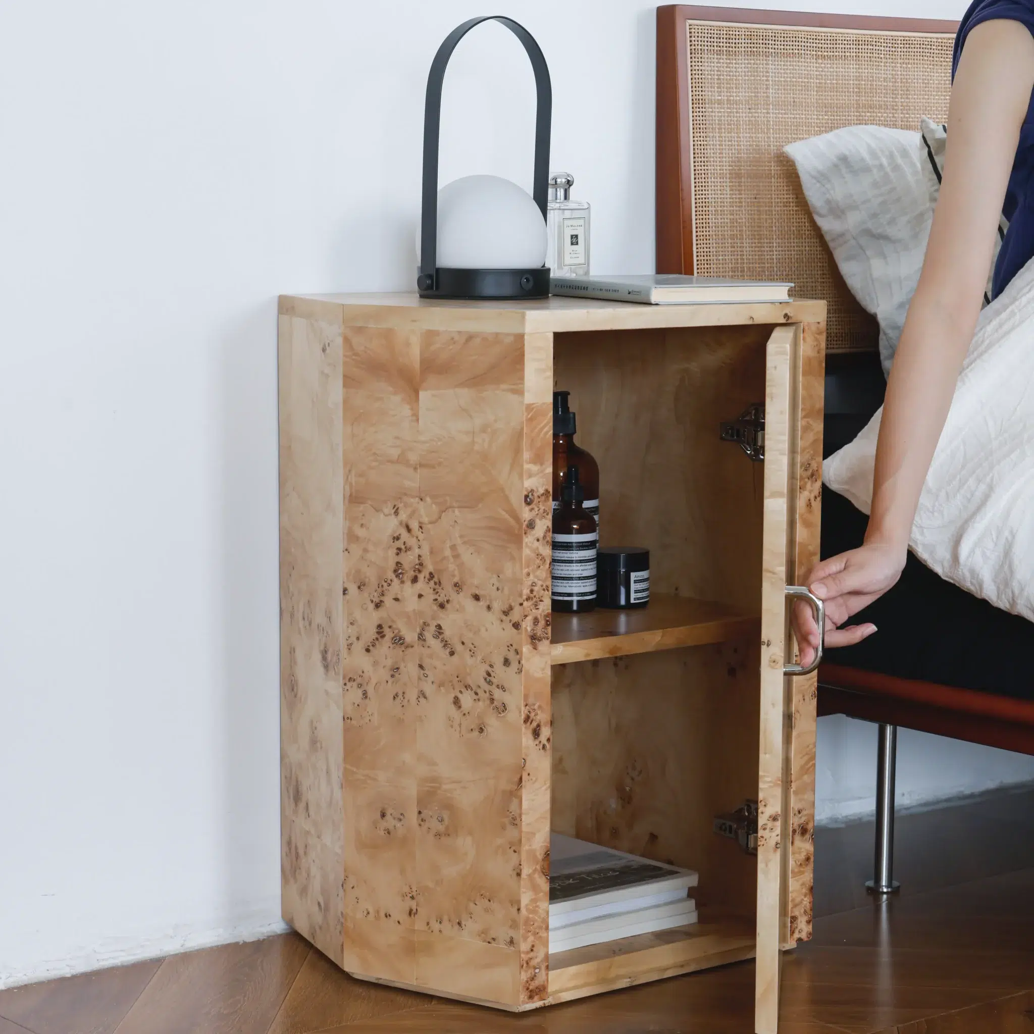 A person opens the door of a light wood bedside table next to a bed. The bedside table has shelves with bottles and books inside, and a modern lamp, perfume, and small items on top.