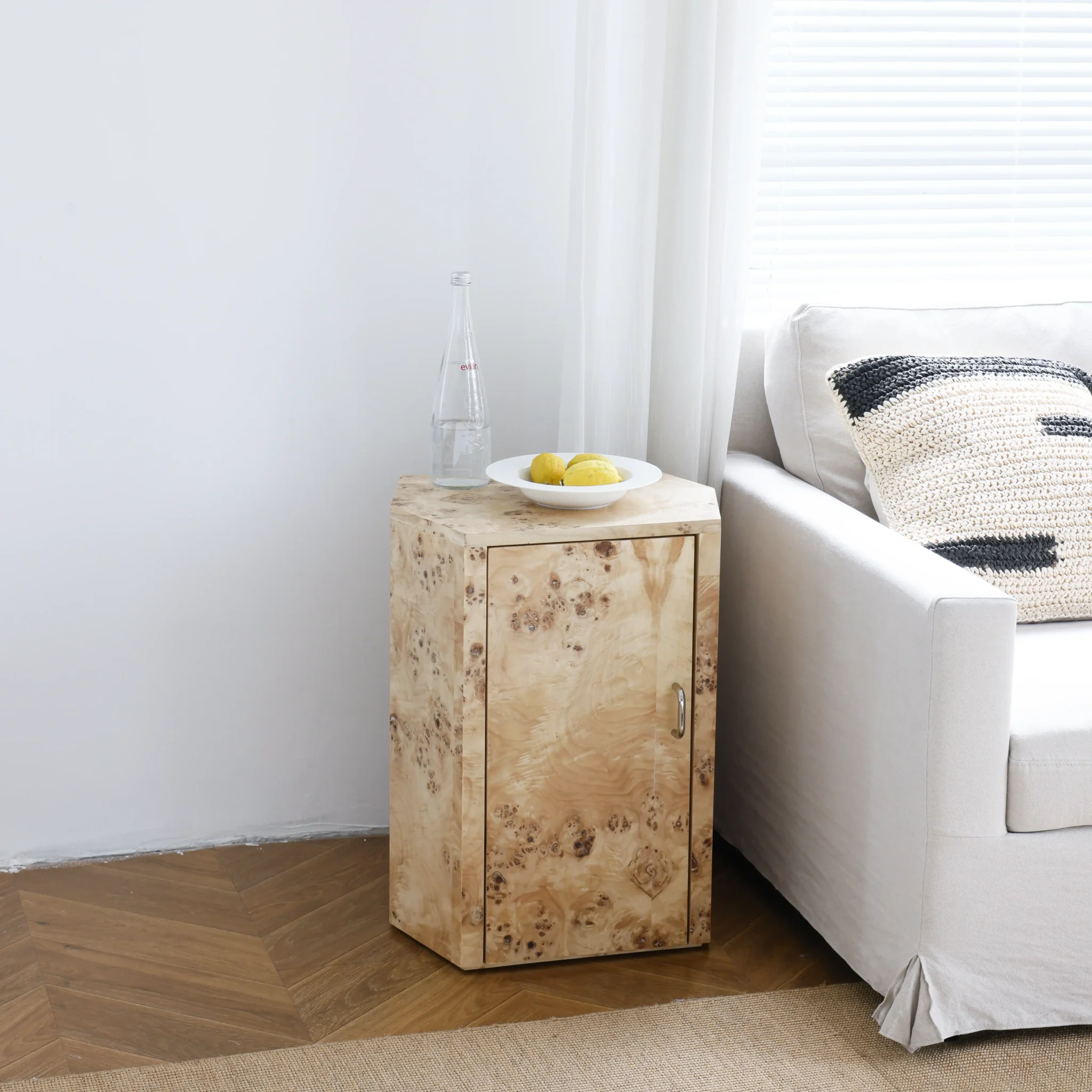 A small wooden side table with a cupboard door stands next to a white sofa. On top, there is a clear glass bottle and a white plate with yellow lemons. Sunlight streams in through a window with white blinds.