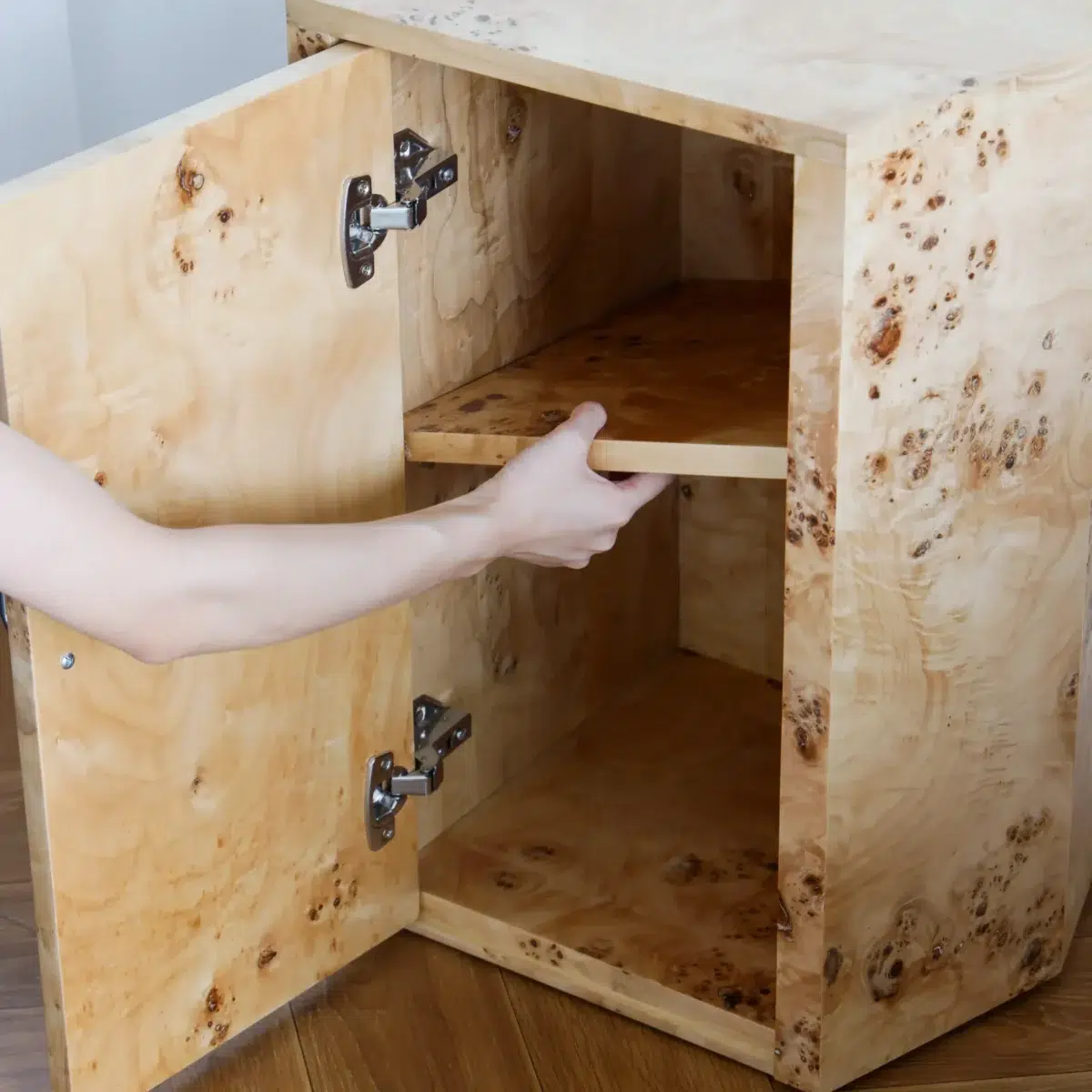 Ptolemy Nightstand_Inside Detail_Front View(4) A person adjusts the removable shelf inside a wooden cupboard with a single door, revealing the interior of the cupboard and its metal hinges.