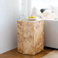 A light wood side table with a natural, burled finish stands beside a beige sofa. On the table are a clear glass bottle, a drinking glass, and a white bowl containing lemons.