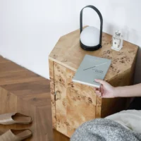 A hand places a book on a wooden bedside table with a lantern-style lamp and a glass perfume bottle. Slippers are on the wooden floor next to the bedside table, beside a bed with a grey blanket.