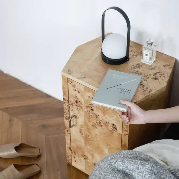 A hand places a book on a wooden bedside table with a lantern-style lamp and a glass perfume bottle. Slippers are on the wooden floor next to the bedside table, beside a bed with a grey blanket.