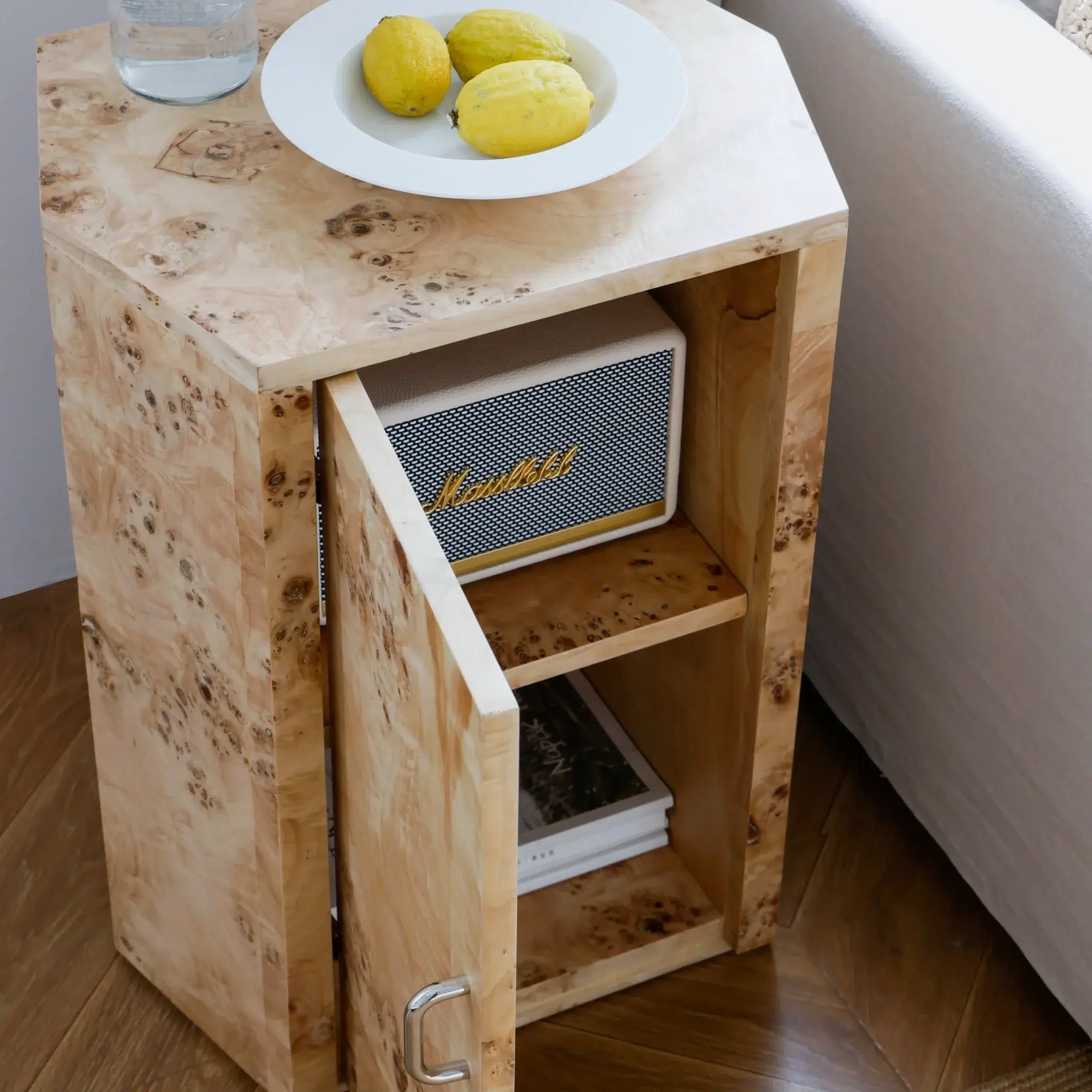A light wood side table with an open door revealing a speaker and books inside. On top, there is a white plate with three lemons and a glass of water, placed next to a white sofa.