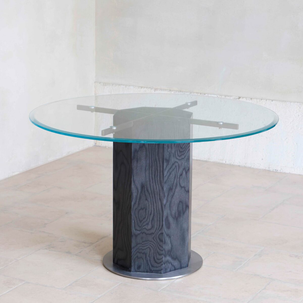 Ptolemy Round Dining Table_Black_C2 A round glass-topped table with a thick, dark wood cylindrical base, set on a light-coloured tiled floor against plain white walls.