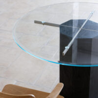 A close-up view of a modern round glass table with a dark geometric base, metal cross support, and a wooden chair nearby, set on a light-coloured tiled floor.