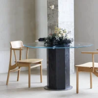 A round glass table with a black cylindrical base is flanked by two light wooden chairs. A floral arrangement with white, yellow, and dark green foliage sits in the centre of the table in a modern, minimalistic room.