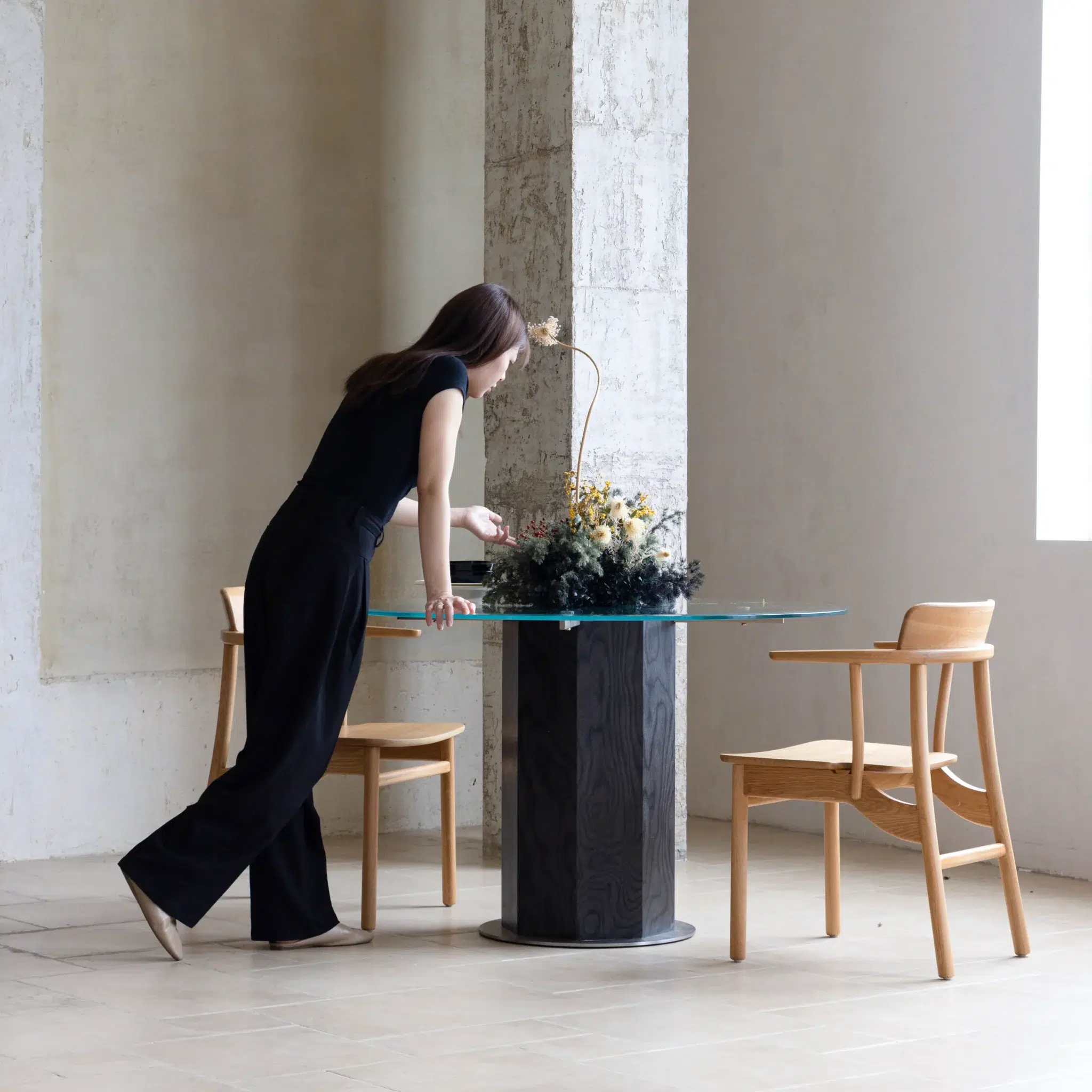 A woman in black clothes leans over a round glass table with a flower arrangement, next to two wooden chairs in a minimalist, light-filled room.