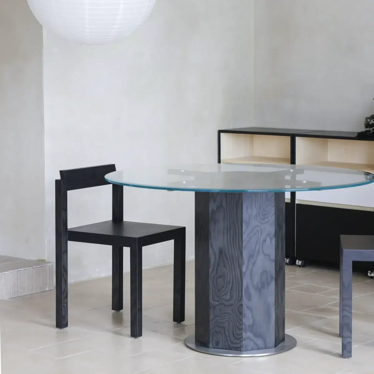Ptolemy Round Dining Table_Black_Front View(2) A modern dining area with a round glass table on a dark cylindrical base, two black wooden chairs, a minimalist shelving unit in the background, and a large white hanging lamp overhead.