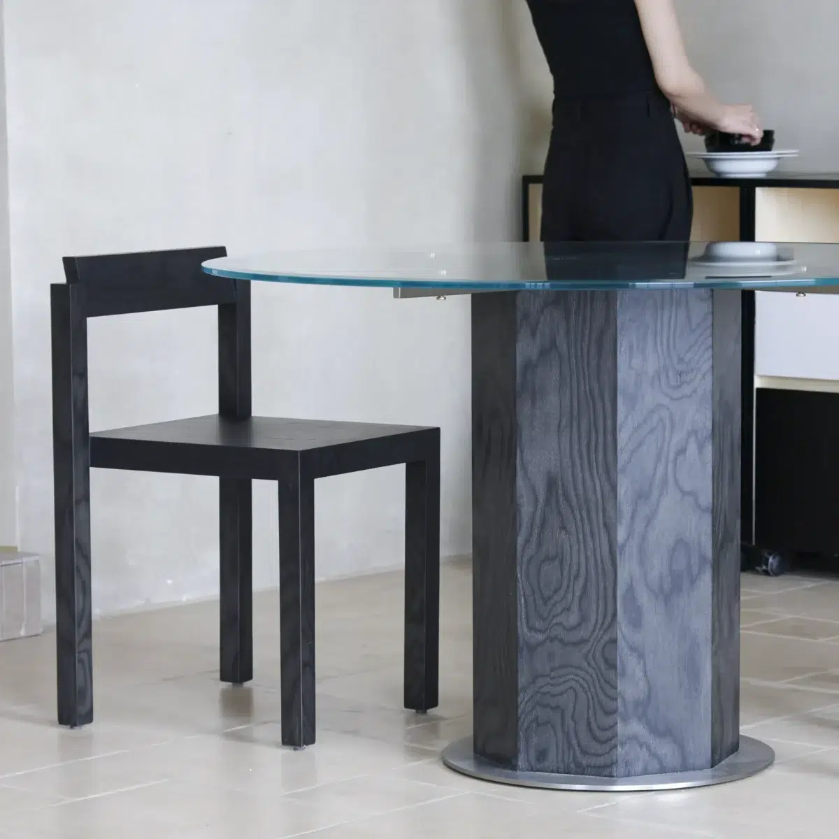 Ptolemy Round Dining Table_Black_Front View(6) A person in black stands near a modern round glass table with a geometric dark wood base and a matching dark wood chair, set against a light-coloured wall and tiled floor.