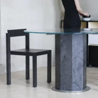 A person in black stands near a modern round glass table with a geometric dark wood base and a matching dark wood chair, set against a light-coloured wall and tiled floor.