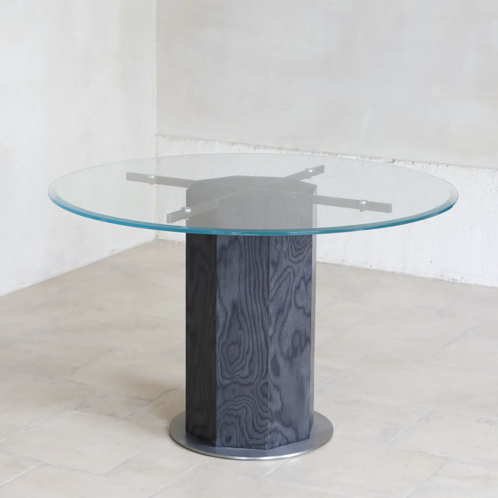 A round glass table with a transparent top and a sturdy, cylindrical black base, positioned in a light-coloured, minimalist room with tiled flooring.