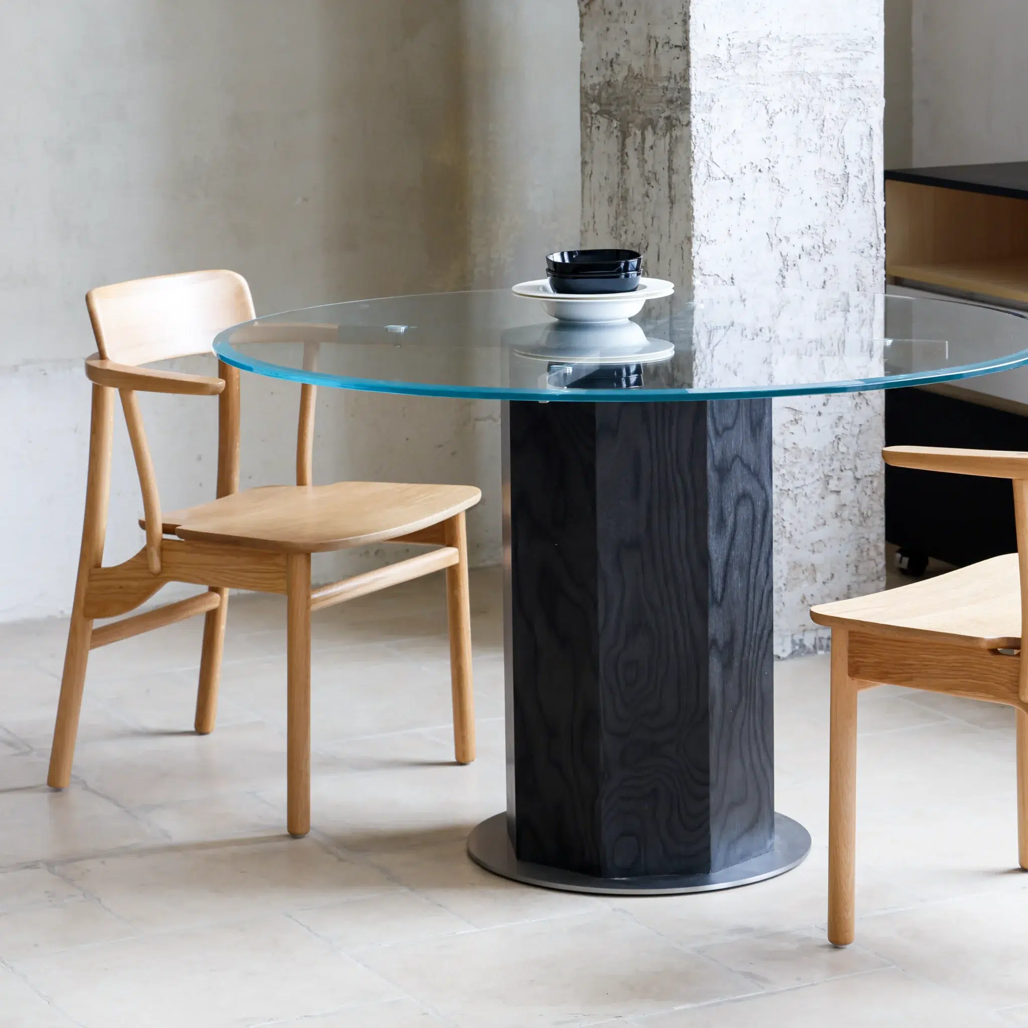 A glass-topped round table with a dark cylindrical base is set with a cup and saucer. Two light wooden chairs are positioned on either side, and the scene has a minimalist, modern interior with concrete and tiled flooring.