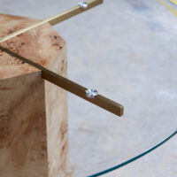 Close-up of a modern table with a round glass top supported by brass metal arms attached to a cylindrical wooden base on a concrete floor.
