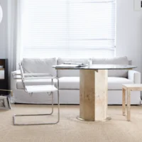 A modern living room with a white sofa, a glass-topped round table with a wooden base, a white metal chair, a light wooden side table, and large window blinds letting in natural light.