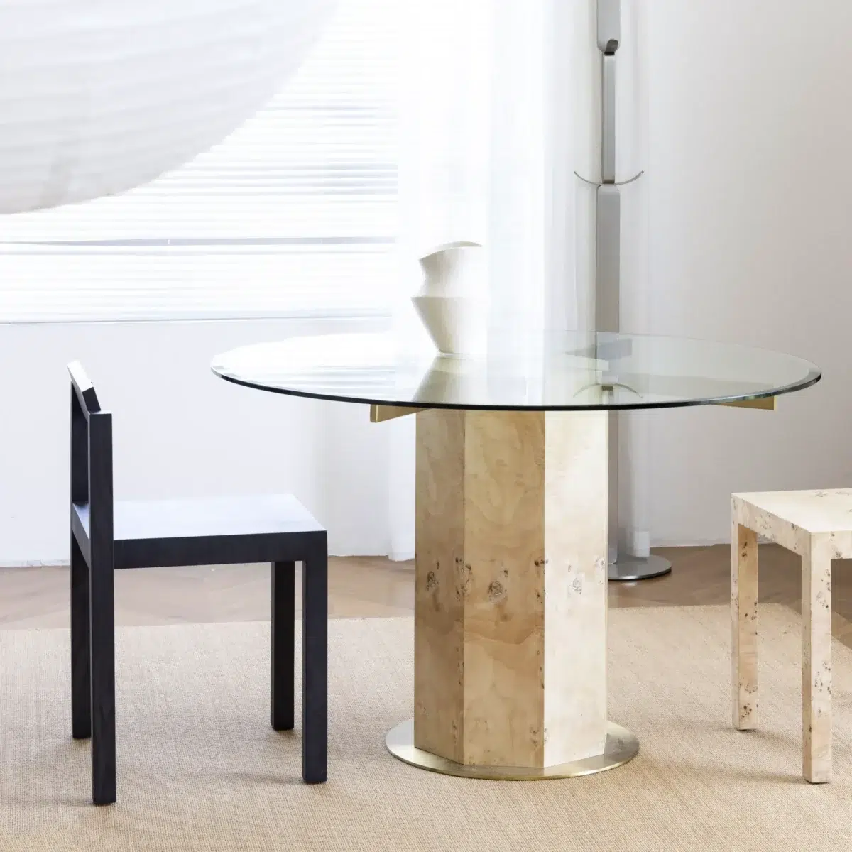 Ptolemy Round Dining Table_Natural_Front View(2) A modern dining area with a round glass table on a light marble pedestal, a black chair, a light stone stool, and a white vase on the table, set on a beige rug with sheer curtains in the background.