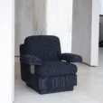 A modern, upholstered armchair with black and dark grey pinstripe fabric sits in a minimalist room with light-coloured stone walls and floors.