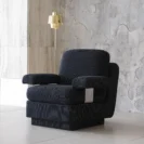 A modern black armchair with textured upholstery sits on a light floor near a concrete wall. A gold floor lamp with a reflective finish stands beside the chair. The setting is minimalist and contemporary.