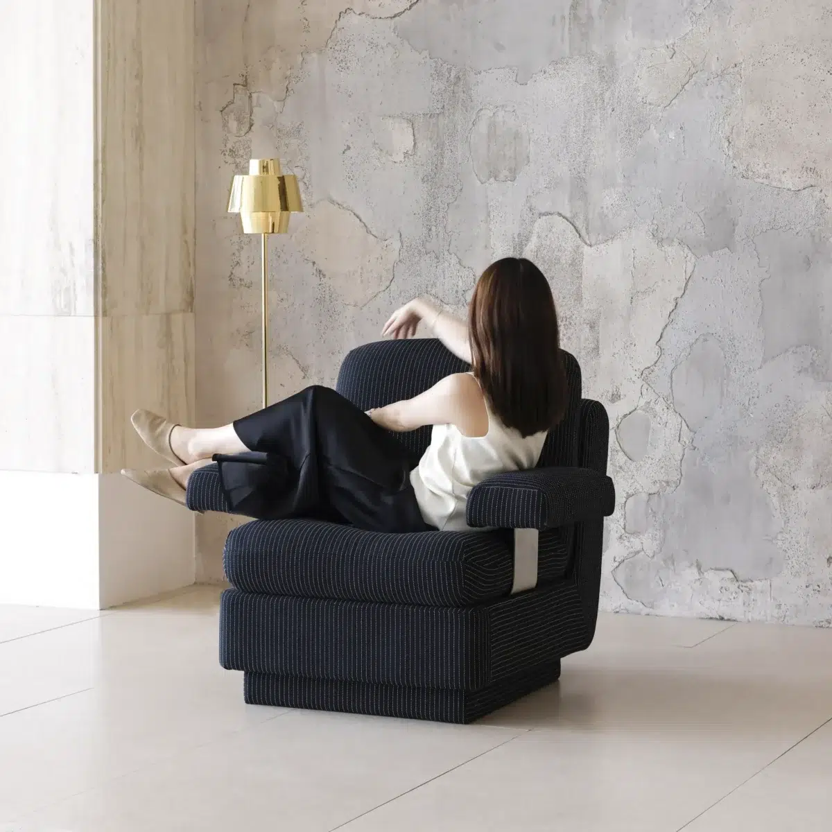 Rest Sofa_Black_Front Side View(8) A woman sits casually in a large, dark armchair with her legs up, facing away. She wears a white top and black trousers. A gold standard lamp stands beside her, against a textured grey wall.