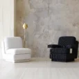 A modern minimalist room with a white armless chair, a black armchair with white detail, and a gold floor lamp. The background features a textured concrete wall and light-coloured tiled floor.