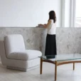 A woman stands at a stone worktop with books and a cup, wearing a white top and long black skirt. In the foreground, there is a modern white armchair and a glass coffee table with wooden legs. The room is bright and minimalistic.