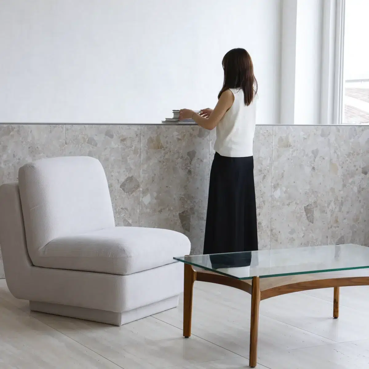 Rest Sofa_White_Front Side View A woman stands at a stone worktop with books and a cup, wearing a white top and long black skirt. In the foreground, there is a modern white armchair and a glass coffee table with wooden legs. The room is bright and minimalistic.