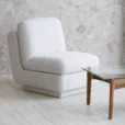 A modern, armless white upholstered chair sits on a light-coloured floor next to a glass coffee table with wooden legs, set against a beige stone wall.