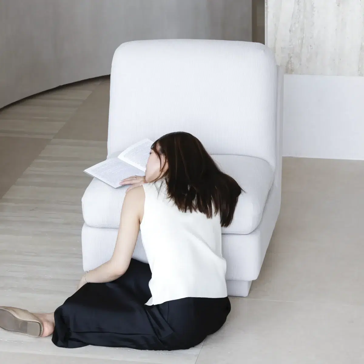 Rest Sofa_White_Front View(2) A woman sits on the floor, leaning against a white chair with her head resting on the seat and her face partially covered by an open book. She wears a sleeveless white top and a long black skirt.