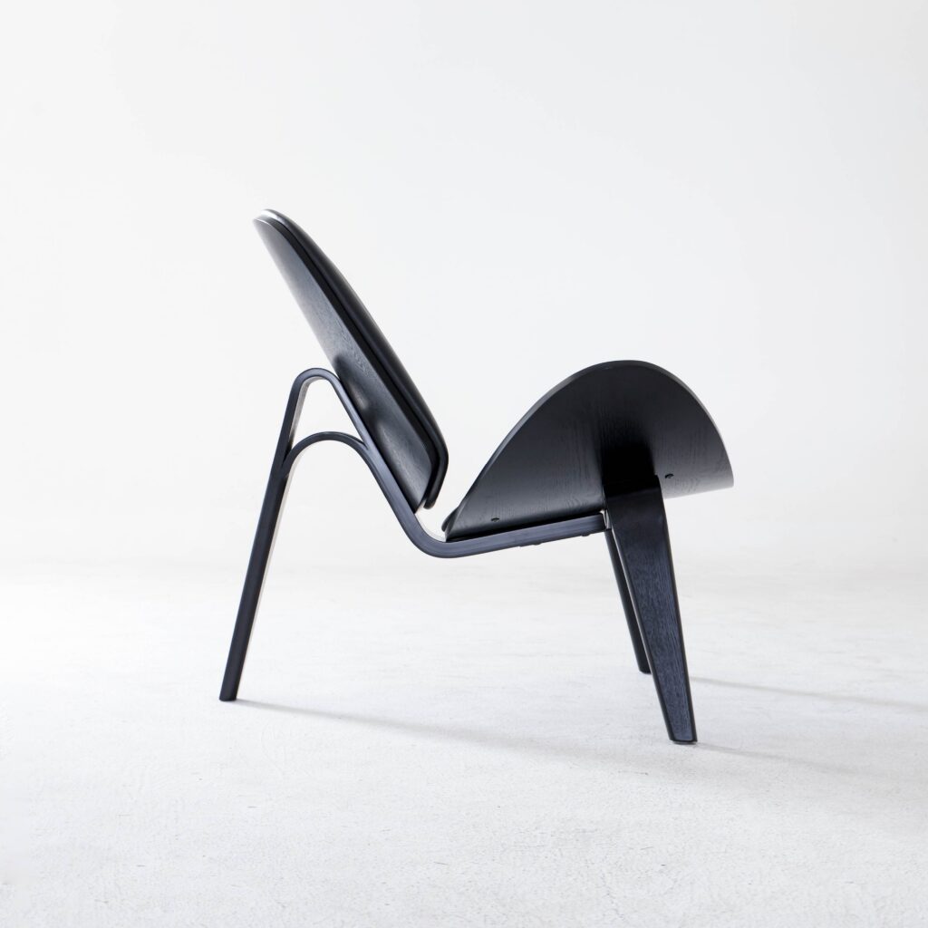 A modern, black, three-legged lounge chair with a curved seat and backrest sits on a light grey floor against a plain white background. The chair has a minimalist and sculptural design.