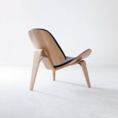 A modern, minimalist wooden lounge chair with curved legs and backrest, featuring a black cushioned seat, is placed in a bright, empty room with white walls and floor.