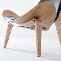 A close-up view of a modern wooden lounge chair with curved legs and a black cushioned seat, set on a light-coloured floor against a plain background.