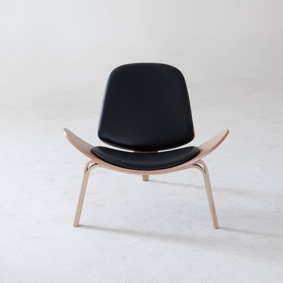 Shell Chair_Natural_B6 A modern lounge chair with a curved wooden frame and black leather seat and backrest, placed on a smooth, light grey floor against a plain white background.