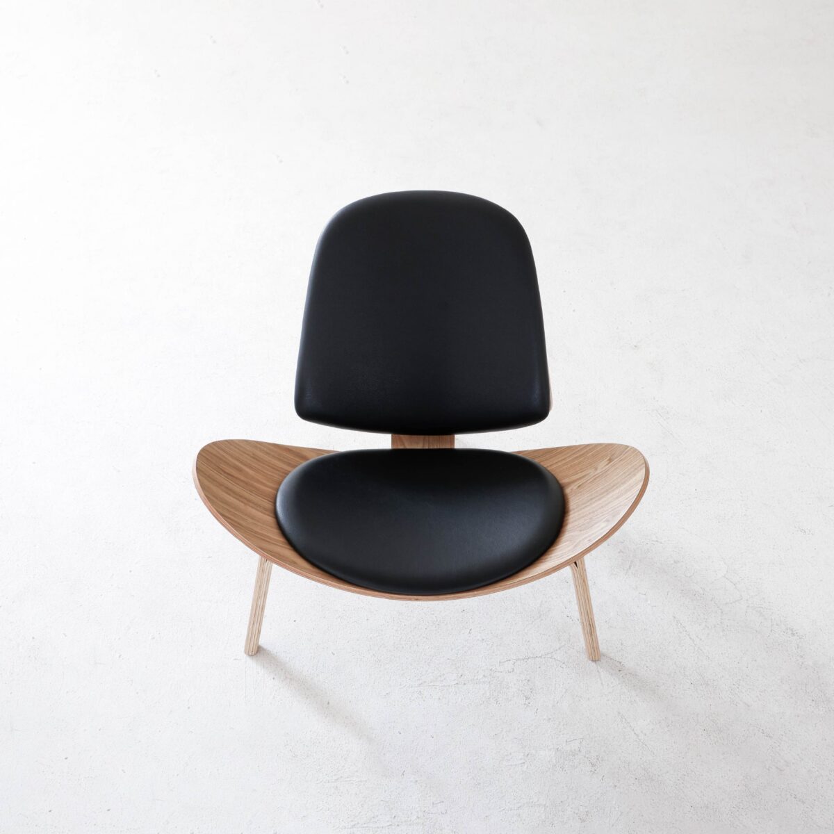 Shell Chair_Natural_B8 Minimalist chair with a curved wooden seat and backrest, both padded with black cushions, photographed from above on a light, textured surface.