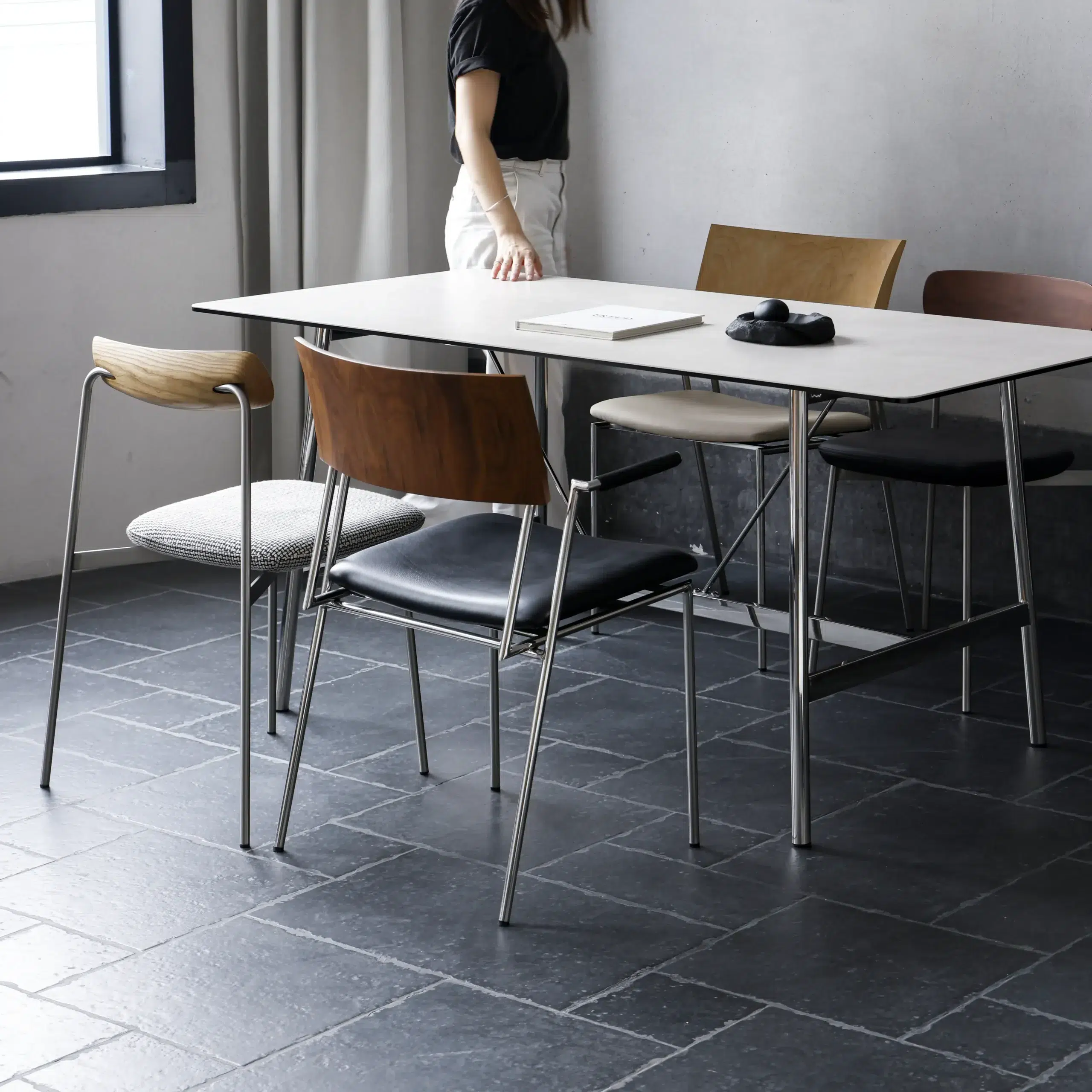 A woman stands near a modern dining table with four mismatched chairs on a dark tiled floor. On the table are a pair of headphones and a closed notebook. Light comes in from a window on the left.