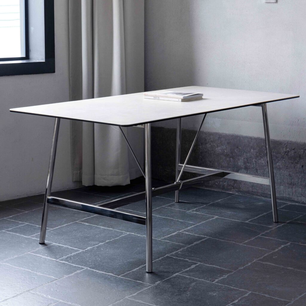 A minimalist table with a thin, light-coloured tabletop and metal legs stands on a dark tiled floor next to a window with light curtains. A closed book or magazine lies on the table.