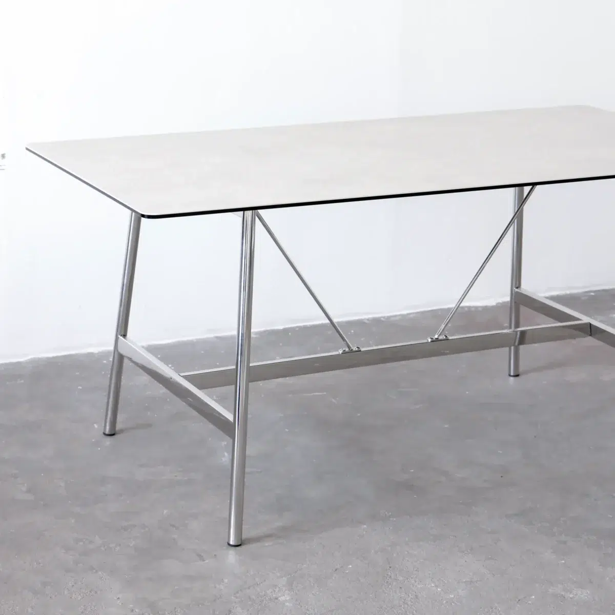 Shuge Dining Table_White_Front View A rectangular table with a thin, light-coloured top and metal legs stands on a grey concrete floor against a white wall. The table features simple, modern design with metal crossbars for added support.
