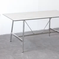 A rectangular table with a thin, light-coloured top and metal legs stands on a grey concrete floor against a white wall. The table features simple, modern design with metal crossbars for added support.